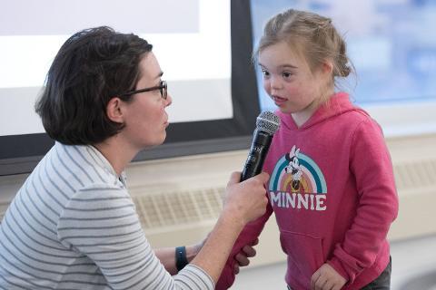 An adult encourages a child with Down syndrome to speak into a microphone during a presentation
