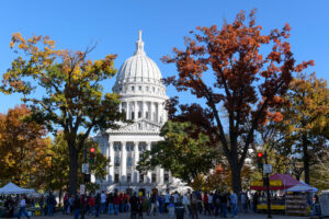 The capitol building peaks through colorful trees, while a crowd of people move down the sidewalk