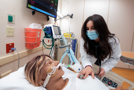 A health care professional practices using their stethoscope on a manikin in a simulation room.