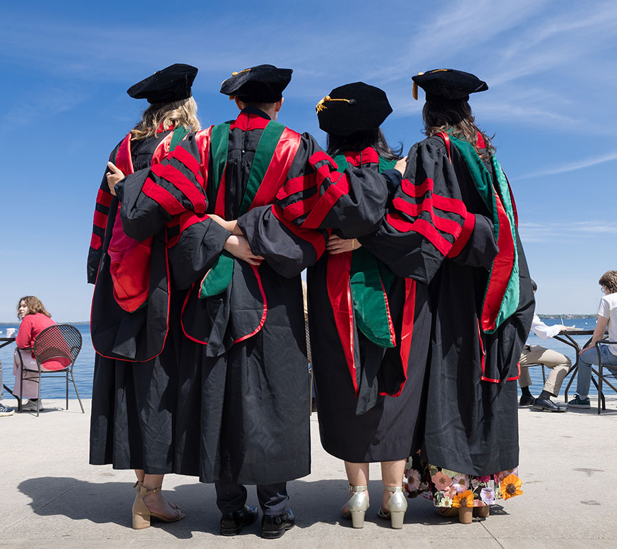 Four MD students in caps and gowns stand arm in arm at the UW Memorial Union Terrace after the graduation ceremony.