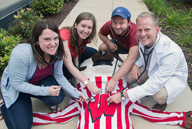 Students holding their stethoscopes to Bucky Badger's chest