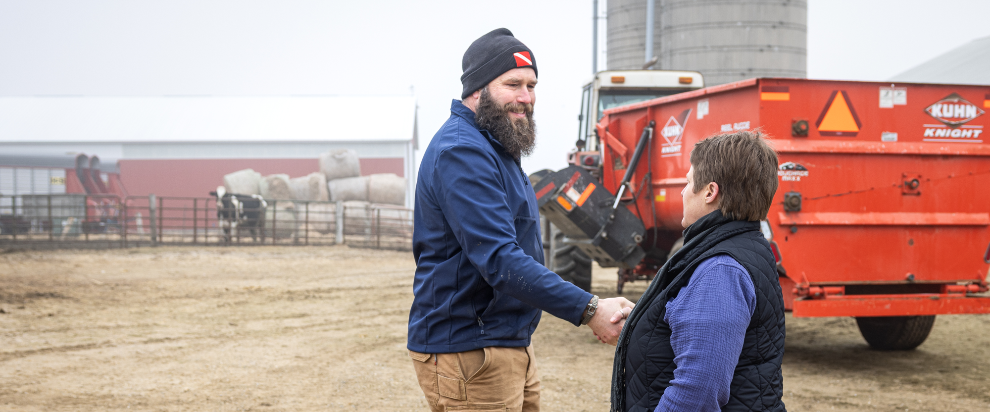 Two people shake hands on a farm near machinery and cows