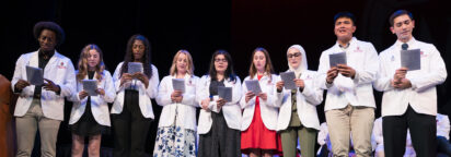 Students in white coats read together on a stage