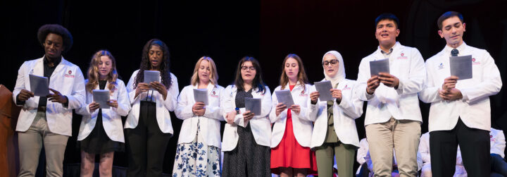 Students in white coats read together on a stage
