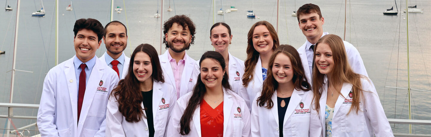 A group of students in white coats smiling together in front of Lake Mendota