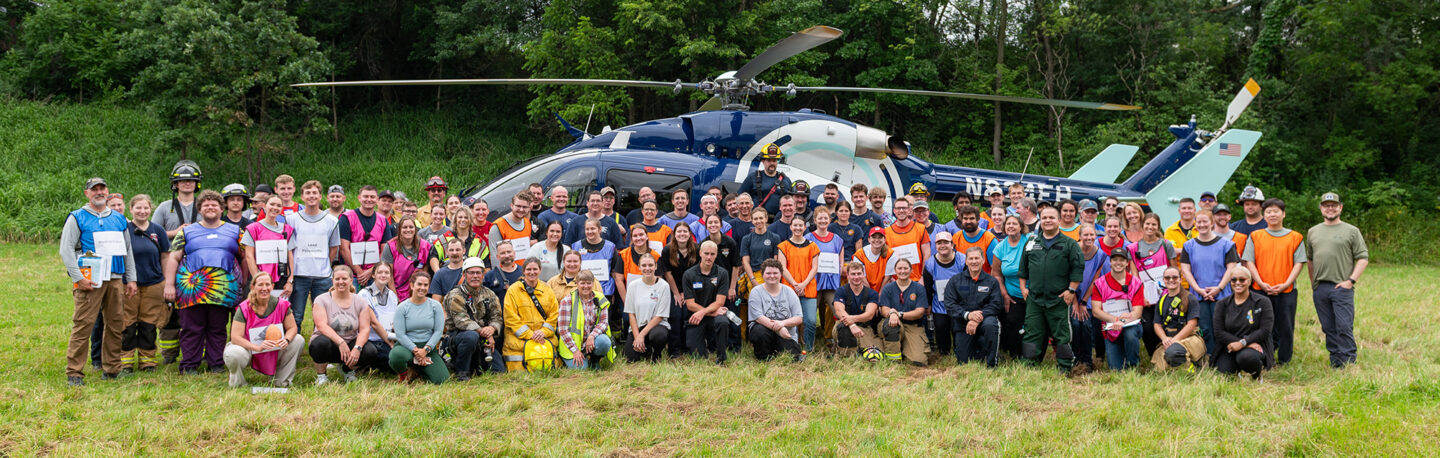 A large group of students and instructors smiling together in front of a rescue helicopter