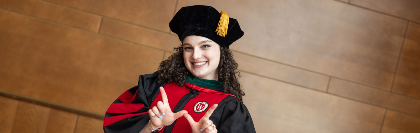 A woman stands in graduation attire and makes a W with their hands