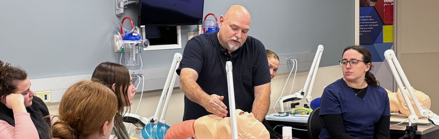 A group of students observe a man demonstrating on a mannequin in a class setting