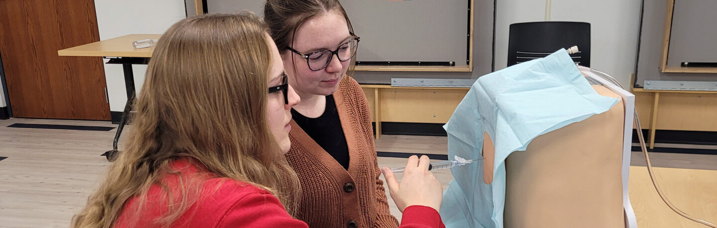 Two physician assistant students working in a skills lab, one practices administering a shot on a manikin